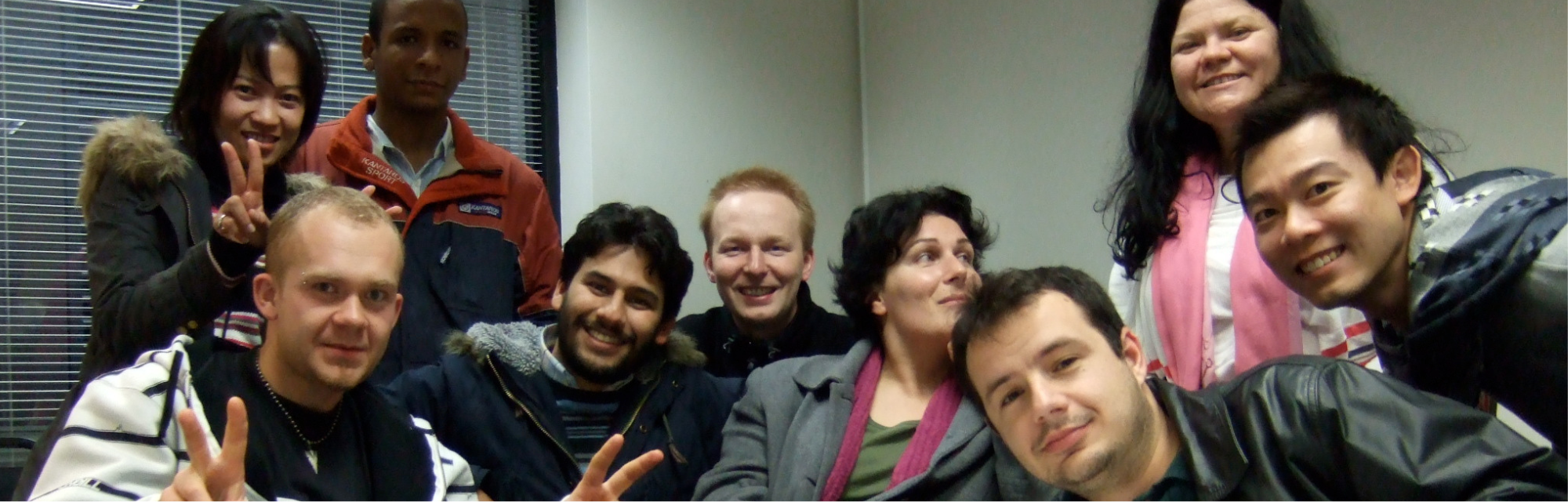 Group of nine diverse people smiling and posing for a photo in a room, some making peace signs.
