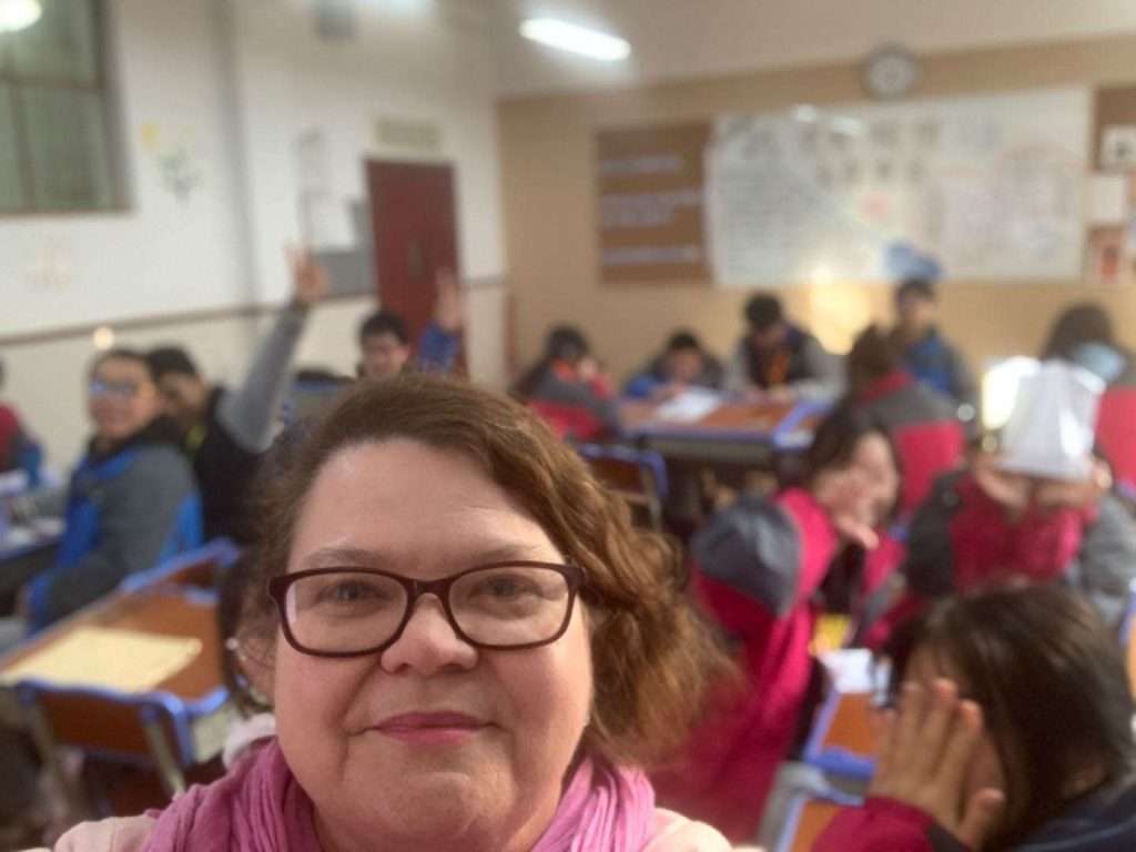 A woman wearing glasses and a pink scarf takes a selfie with a classroom of students in the background, some of whom are raising their hands. The classroom is brightly lit.
